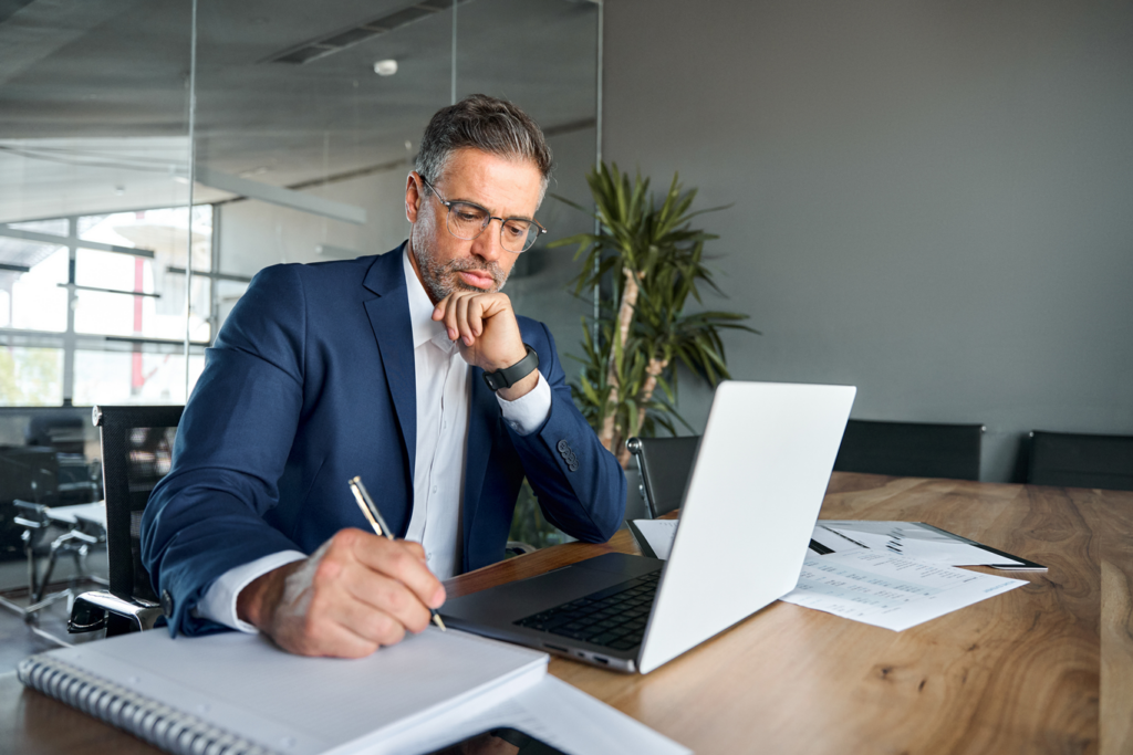 Busy professional middle-aged executive sitting at a desk working on laptop writing notes