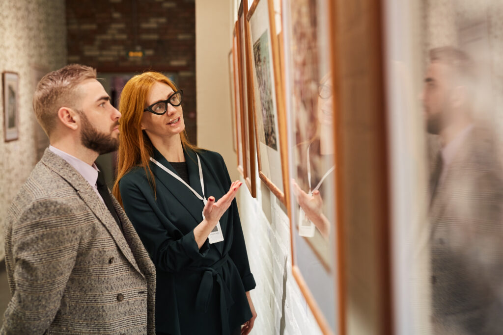 Young art manager pointing at painting and telling about it to young man while they standing at gallery